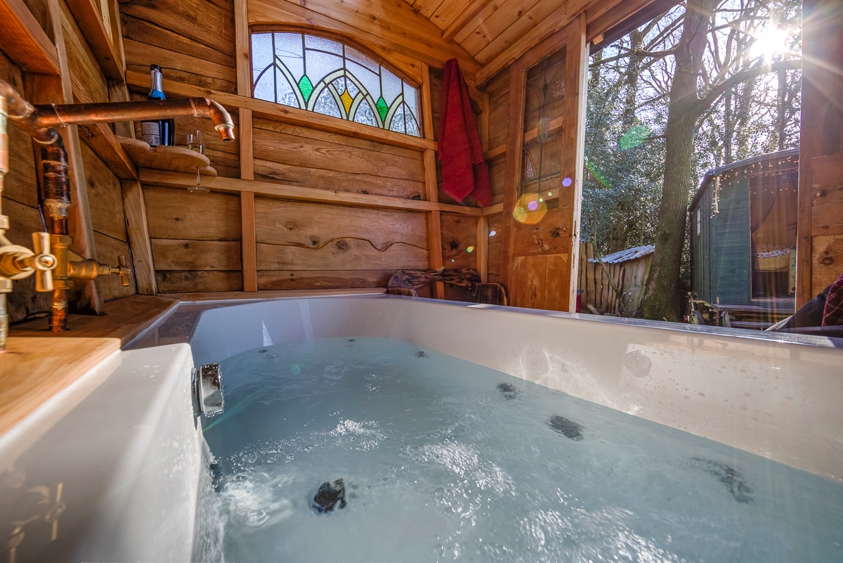 An inviting bathhouse features a spacious double-ended jet bath with clear water, complemented by wooden walls and stained glass accents. Sunlight streams through the windows, illuminating the interior. Storage shelves hold towels and bath products, enhancing the relaxing environment amidst natural surroundings.