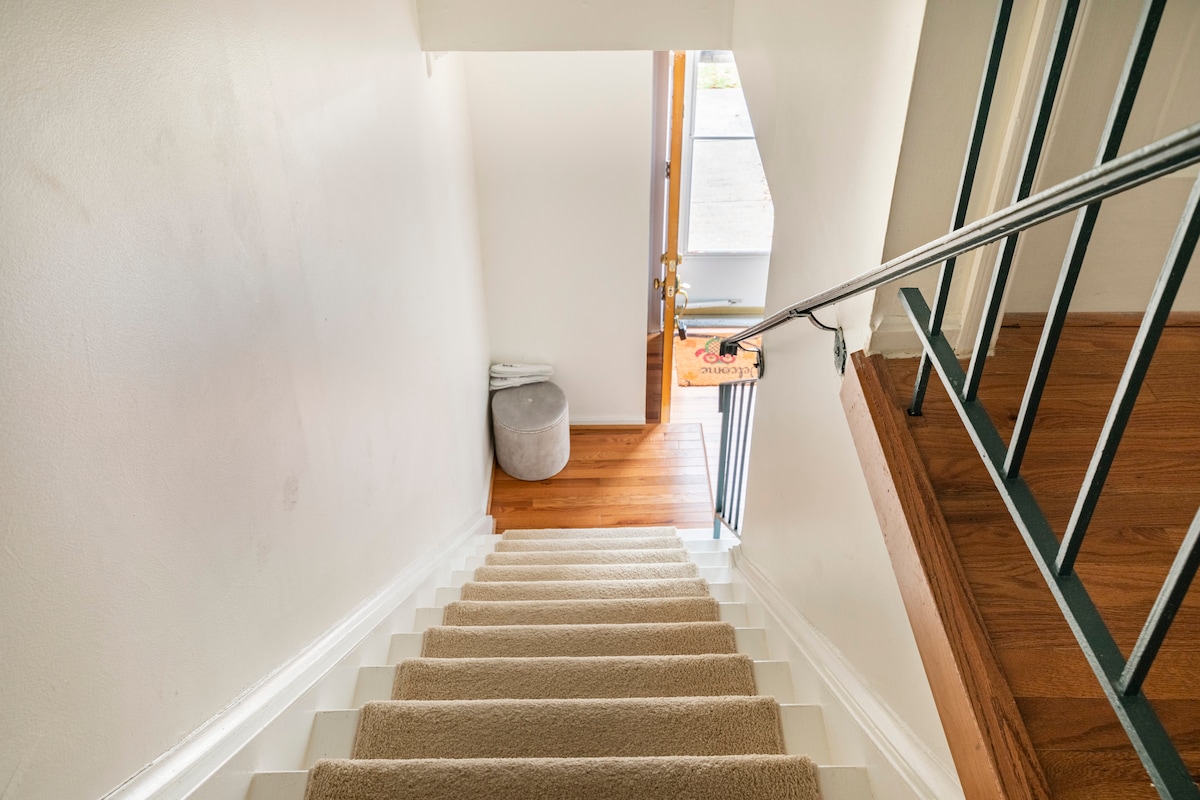 A well-lit staircase leads downwards, featuring a soft carpet runner on the steps. The wooden floor is visible at the base, complementing the neutral wall color. A doorway at the bottom opens to the exterior, hinting at convenient access outside.