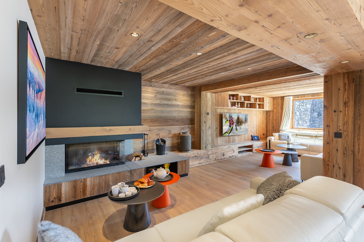 A spacious living room features wooden walls and ceilings, enhancing a rustic aesthetic. A modern fireplace sits between comfortable seating. A television is mounted on one wall, while a coffee table displays snacks and drinks. Natural light filters through large windows.