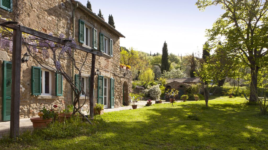 A charming stone villa is set against a backdrop of lush greenery. Sunlight gently illuminates the well-maintained lawn, surrounded by trees and flowering plants. The exterior features traditional shutters and a covered patio, inviting guests to enjoy the tranquil garden atmosphere.