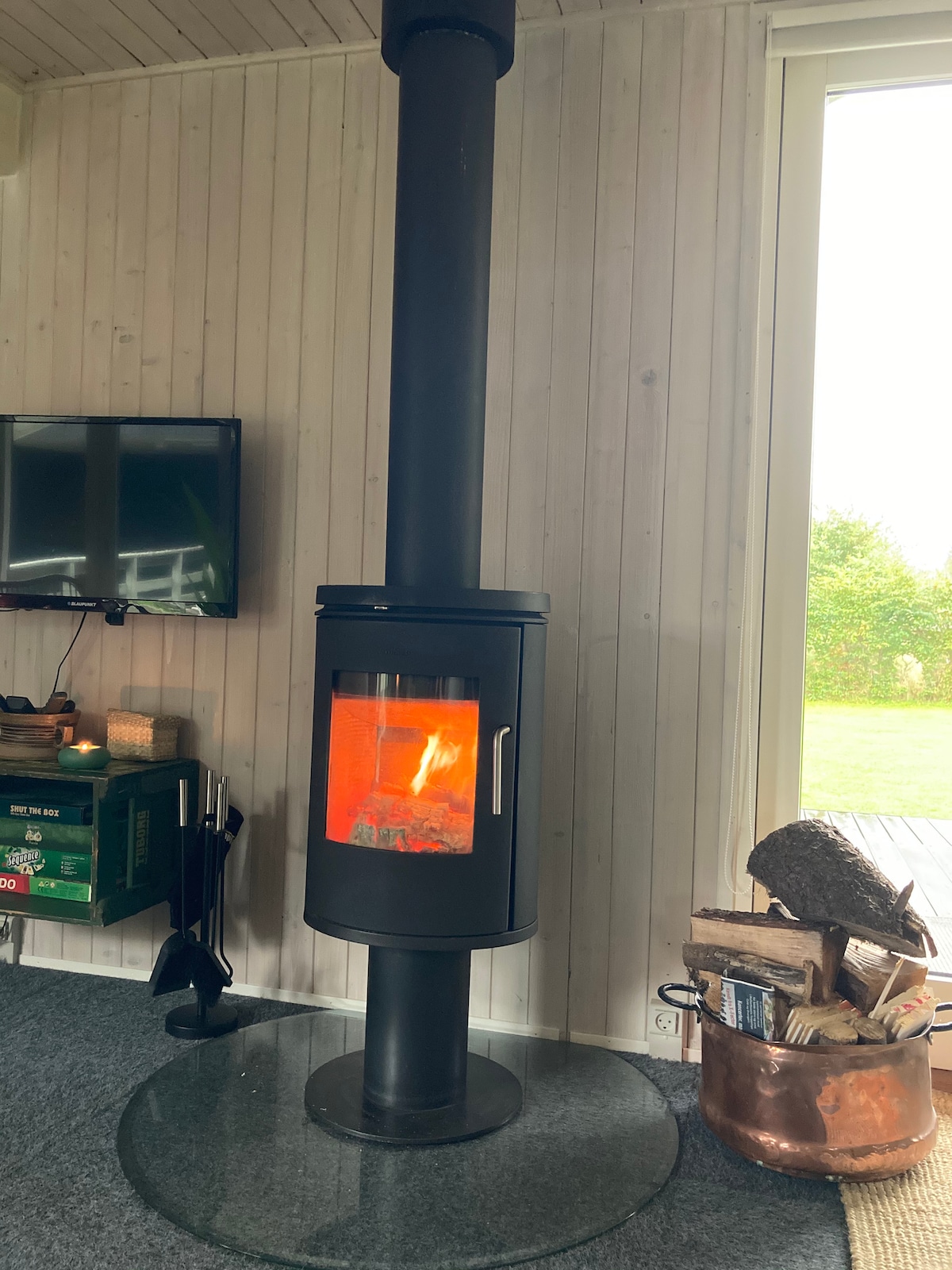 A modern wood-burning stove is positioned on a circular glass base, featuring a glass front that reveals a warm, active flame. A television is mounted on the wall nearby, and natural light enters through a large window, illuminating the area.