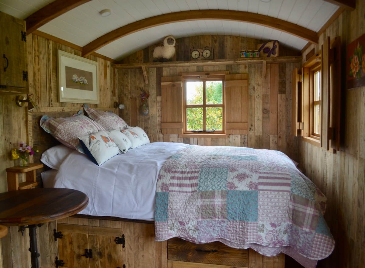 A cozy sleeping area features a spacious bed adorned with a patchwork quilt and decorative pillows. Natural light filters through two wooden windows, complementing the rustic timber walls. A small side table adds functionality to the tranquil space.