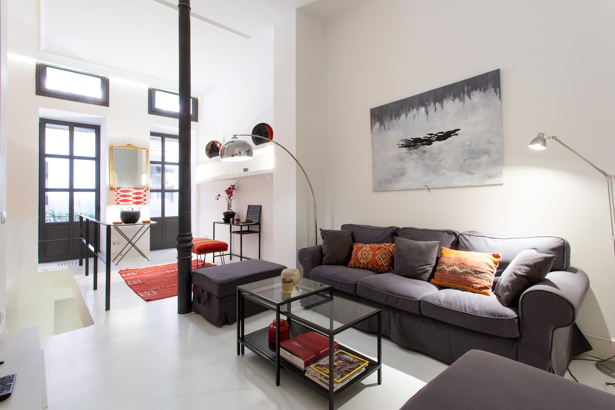 A cozy living area features a gray sofa adorned with decorative pillows and a glass coffee table. Natural light enters through large windows with dark frames. An artistic wall painting and a floor lamp enhance the inviting atmosphere. A dining table and chair are positioned in the background.