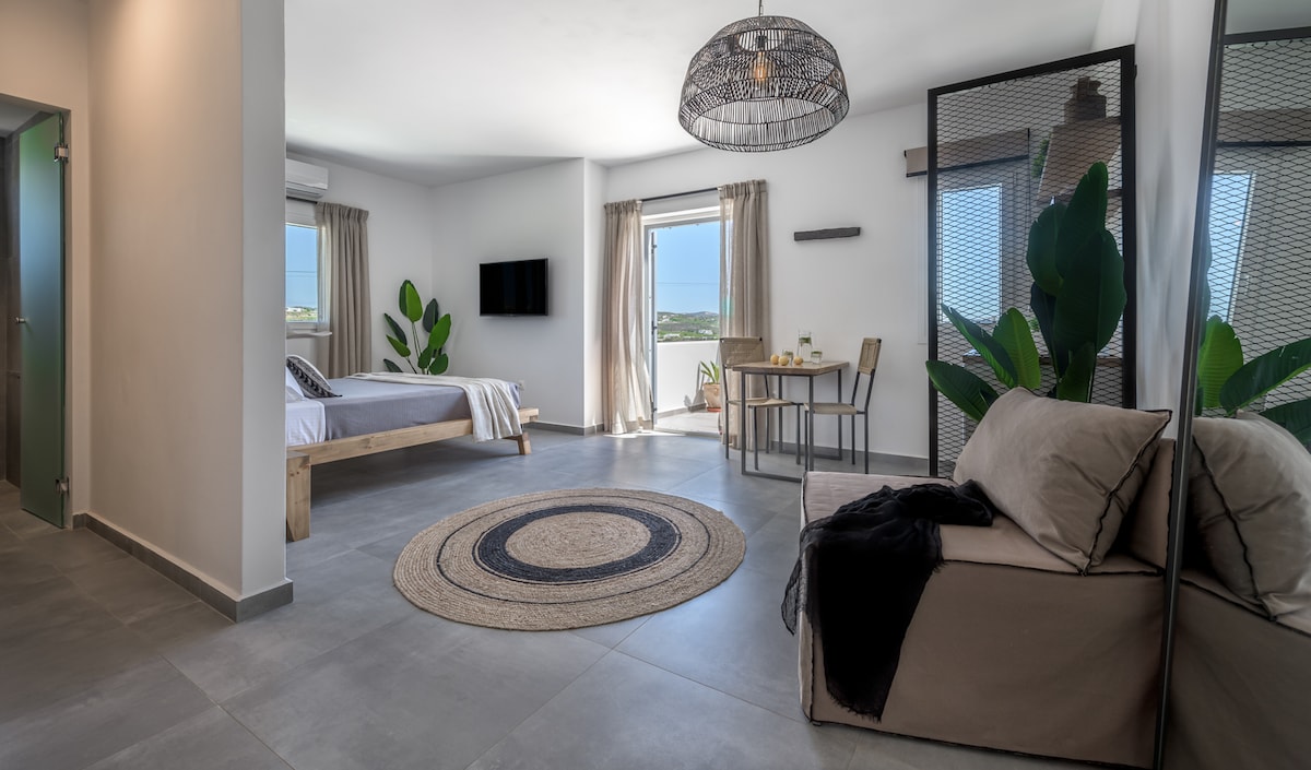The studio space features a queen-sized bed with neutral linens, complemented by large windows that allow natural light to fill the area. A dining table with two chairs is positioned next to a cozy sofa, while green plants provide a touch of nature to the modern decor.