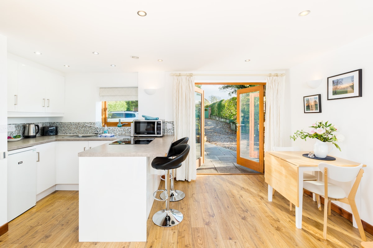 A bright and open kitchen area features modern white cabinetry and a large countertop with bar seating. Natural light floods the space through French doors that lead to an outdoor terrace. A dining table is positioned against the wall, accompanied by chairs.
