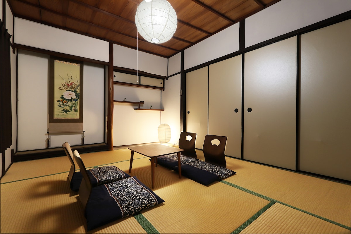 A traditional Japanese room features tatami mat flooring and low seating with decorative cushions. A wooden table sits at the center, illuminated by a round paper lantern hanging overhead. Sliding shoji doors are partially closed, showcasing a serene wall with floral artwork.