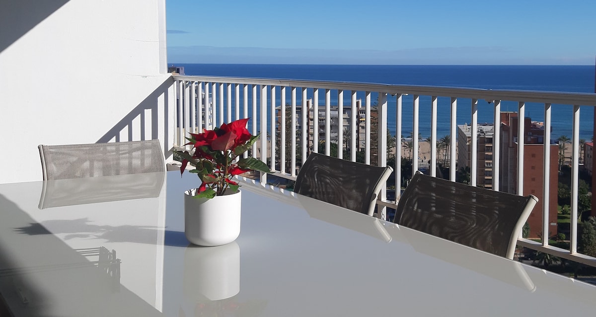 A balcony table is set with a white planter containing a red flower, overlooking a clear blue sea. The scene includes sleek, light-colored chairs around the table, framed by a railing that enhances the view of the coastline and horizon.
