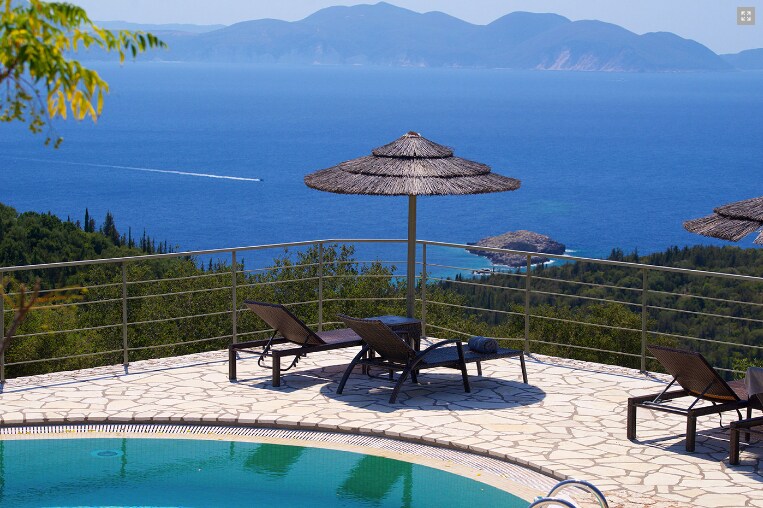 A tranquil swimming pool area features sun loungers and umbrellas, offering a stunning view of the Ionian Sea and distant islands. The stone terrace complements the surroundings, creating a serene environment for relaxation and enjoyment of the landscape.