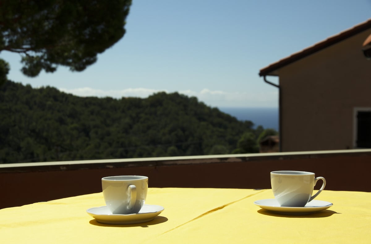 Two white cups are positioned on saucers atop a yellow tablecloth, with a scenic view of a green hillside and the sea in the background. The clear sky enhances the inviting atmosphere of the terrace.
