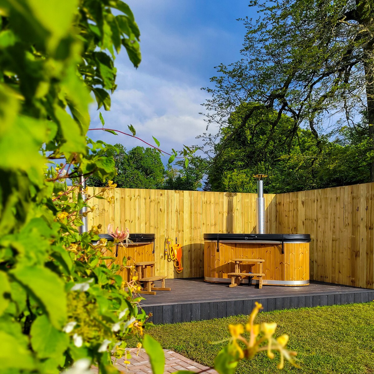 Two wood-fired hot tubs are situated on a wooden deck surrounded by a fenced area. Each tub features a small bench nearby and is framed by greenery, highlighting the outdoor relaxation space.