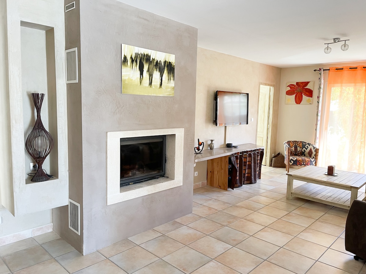 An inviting living area features a modern fireplace integrated into a textured wall. A flat-screen television is mounted above a wooden console, while colorful artwork and plush seating add character. Natural light flows through large windows, complementing the tiled floor.