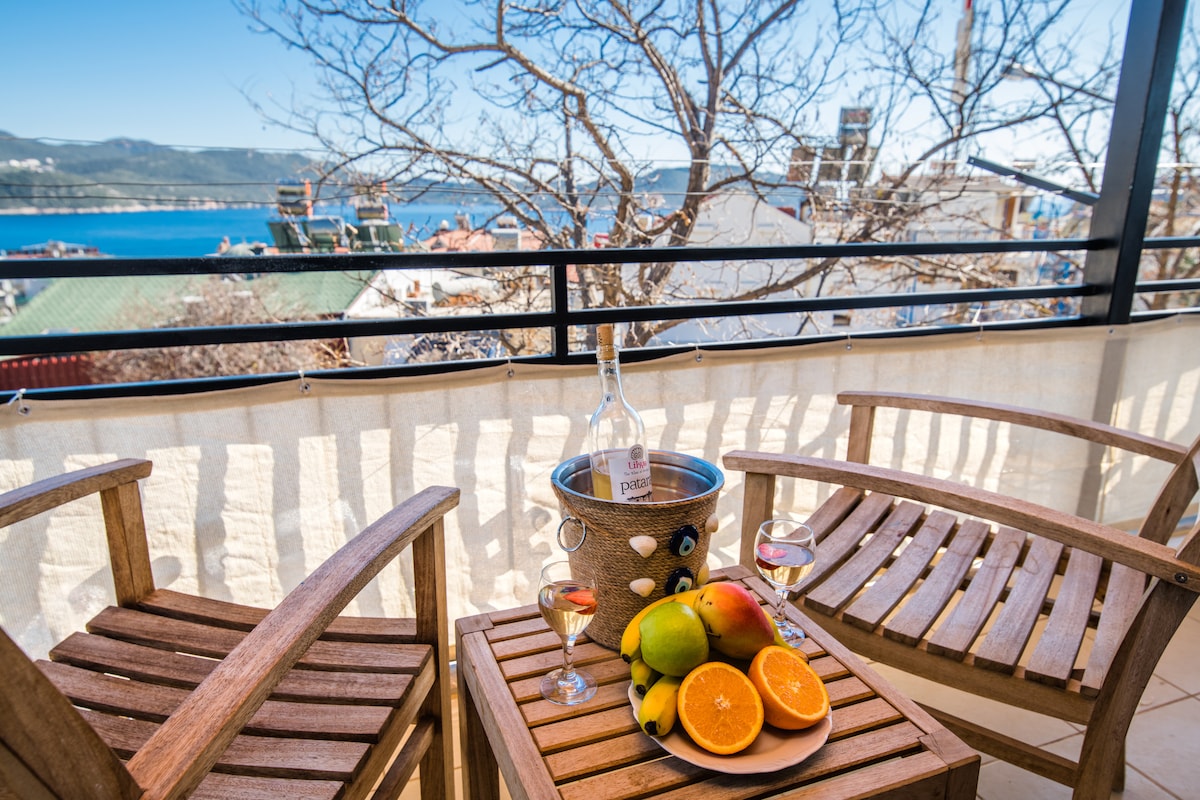 A balcony furnished with two wooden chairs and a small table. A bottle of wine is placed in an ice bucket, accompanied by two glasses and a colorful assortment of fruits, including oranges and mangoes. The background features a scenic view of the sea and distant hills.