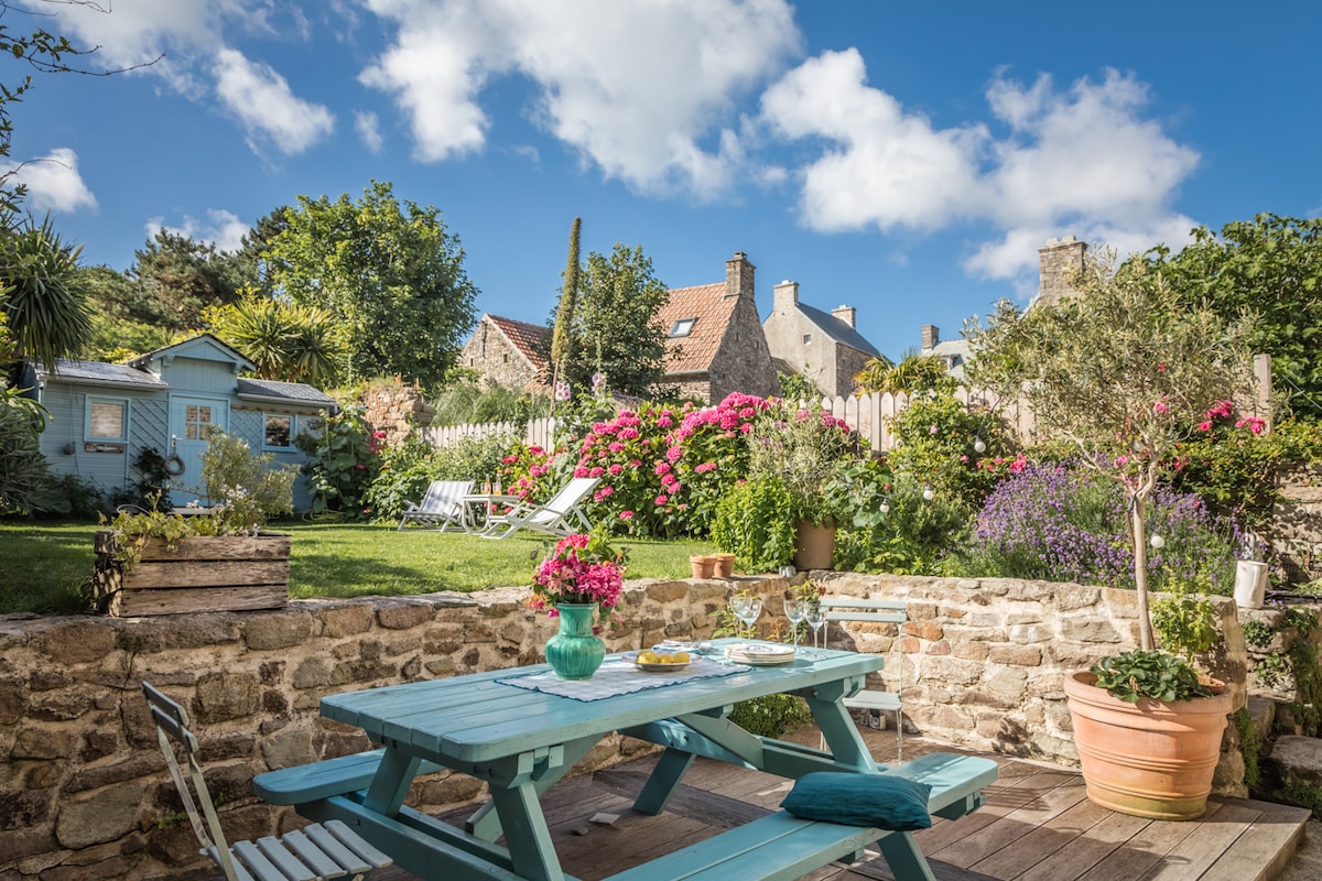 A charming outdoor space features a wooden table set with dishes. Surrounding flowers in various colors bloom vibrantly, with green foliage providing a lush backdrop. A light blue garden shed is visible, along with sun loungers placed on the grass under a partly cloudy sky.