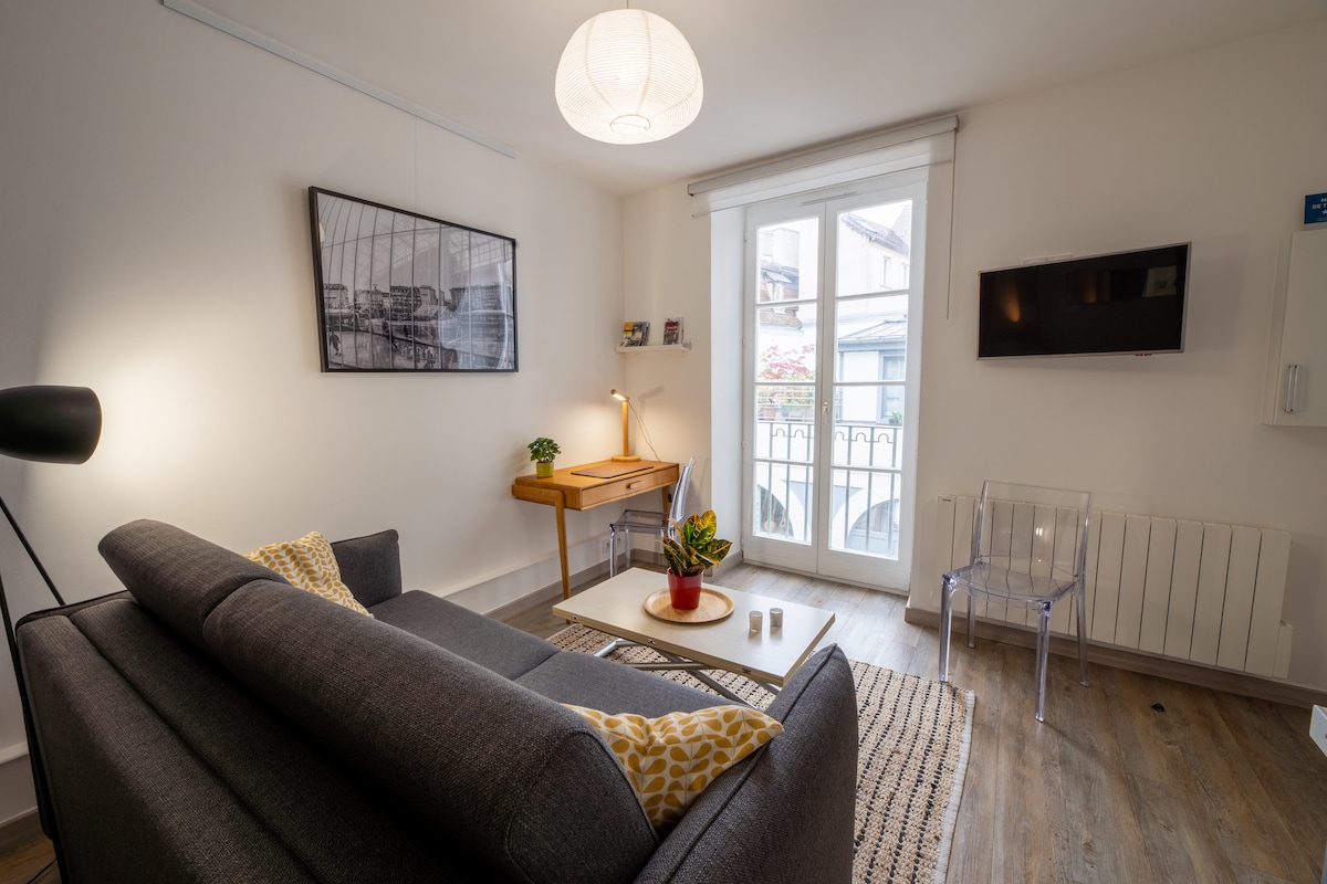 A bright living area features a comfortable gray sofa and a modern wooden desk. Natural light flows through glass doors that open to a small balcony, while a flat-screen television is mounted on the wall. A decorative plant adds a touch of greenery to the space.