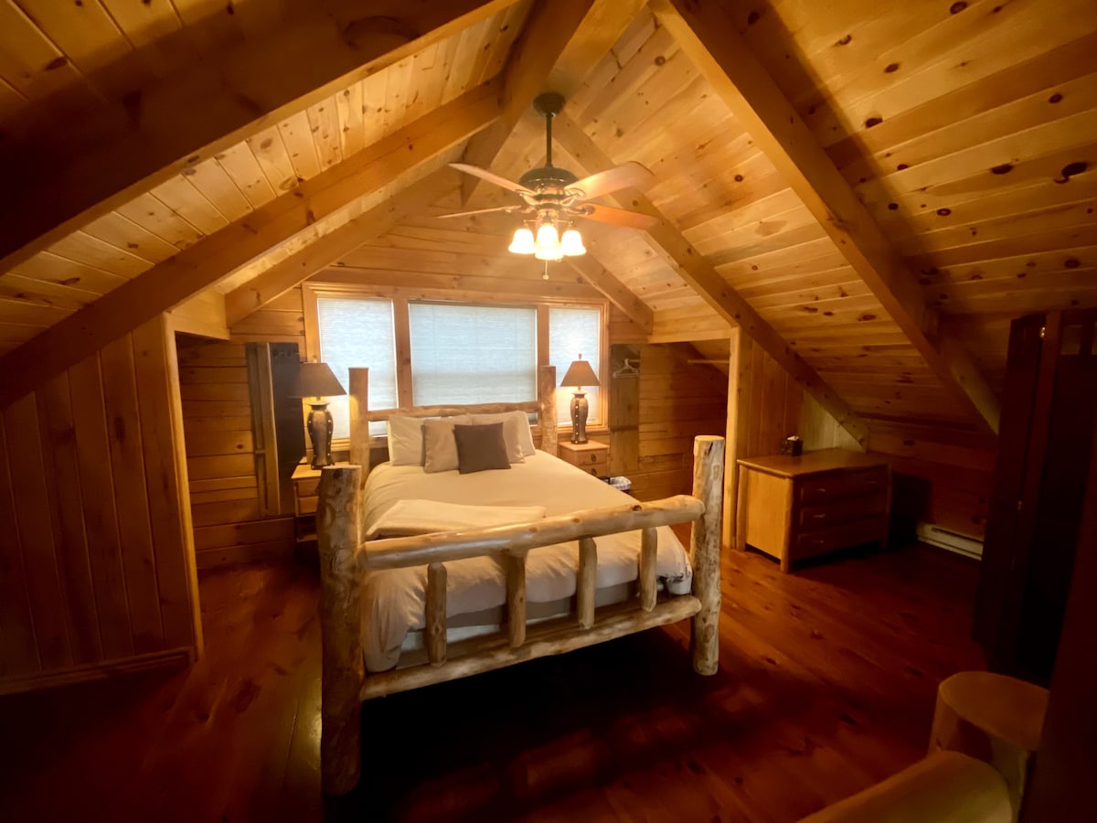 A cozy bedroom features a spacious bed framed with natural wood logs, creating a rustic ambiance. Soft lighting is provided by a ceiling fan and bedside lamps. Large windows allow for natural light to fill the room, enhancing the warmth of the wooden paneling.