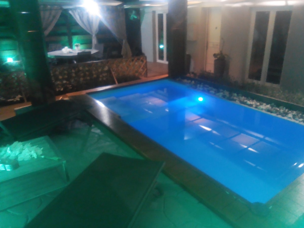 A private pool glows softly under ambient lighting, surrounded by lounge chairs. The pool area is bordered by decorative stones and plants, with a dining area visible in the background, creating a seamless indoor-outdoor connection.