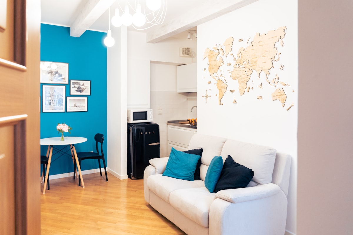 A bright living space showcases a comfortable two-seater sofa adorned with dark blue cushions. A round dining table with black chairs is set beside a light blue wall. A decorative world map is artfully displayed on the adjacent white wall, while a small kitchenette is visible in the background.