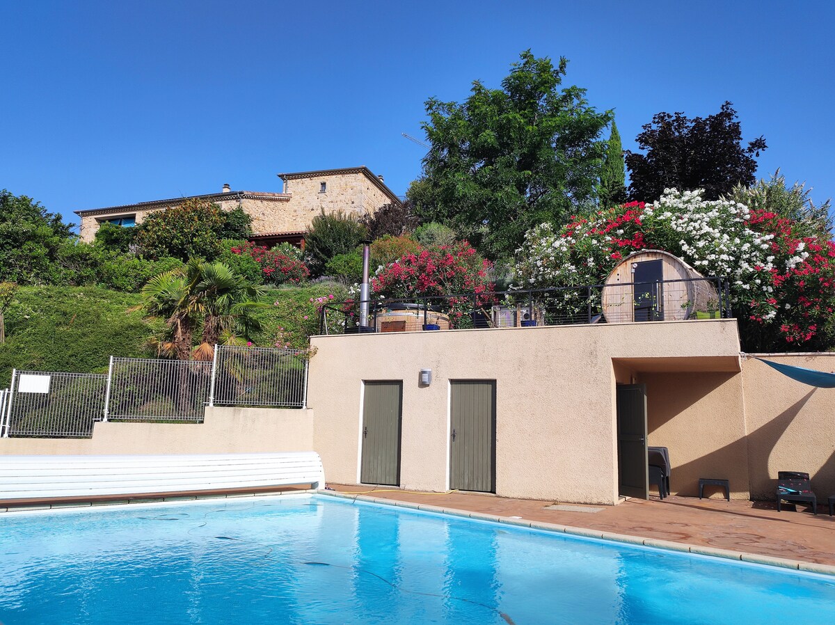 A shared outdoor pool reflects the clear blue sky, bordered by a terrace featuring landscaped gardens. The gîte is visible on the hillside, surrounded by greenery and blooming flowers, offering a serene setting for relaxation.
