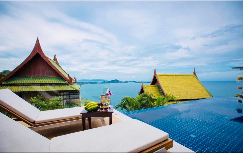 A pair of sun loungers is positioned by an infinity pool, overlooking the ocean. A small table rests between them, featuring a fruit bowl and a bottle of champagne. Traditional Thai roofs are visible in the background, framed by lush greenery under a cloudy sky.