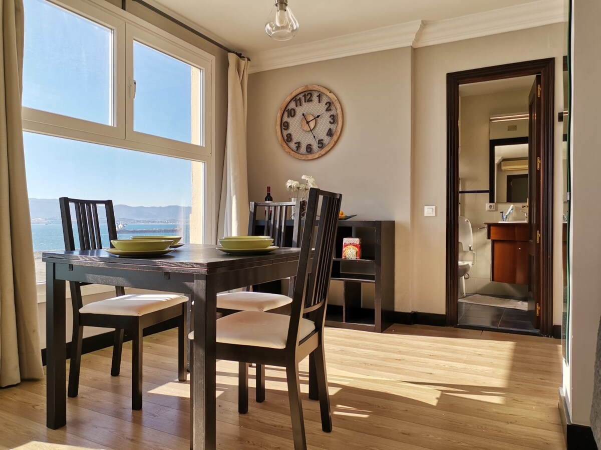 A dining area is featured with a dark wooden table set for four. Large windows provide natural light and views of the sea. A wall clock adds a functional touch, while a small cabinet and visible bathroom doorway complete the space.