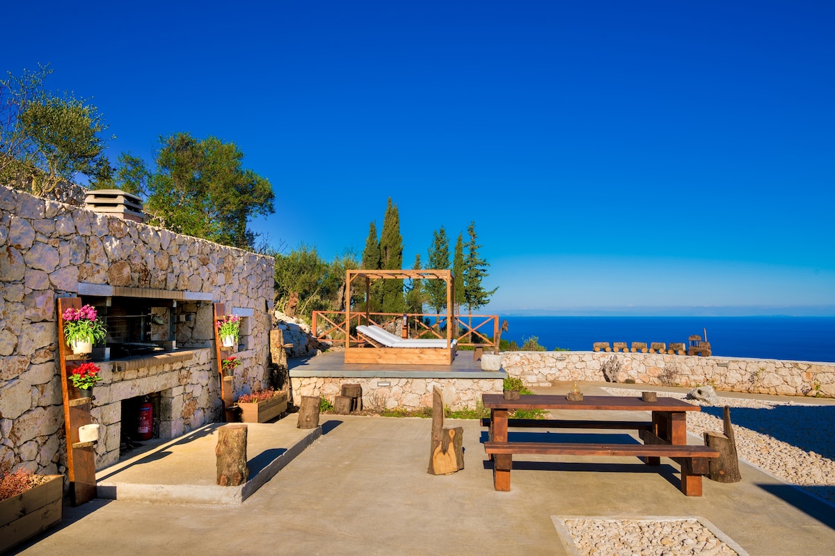 An outdoor area features a stone barbecue station, complemented by a large wooden dining table and benches made from natural materials. A wooden pergola provides shade, while the infinity view of the sea and distant trees enhance the serene atmosphere.