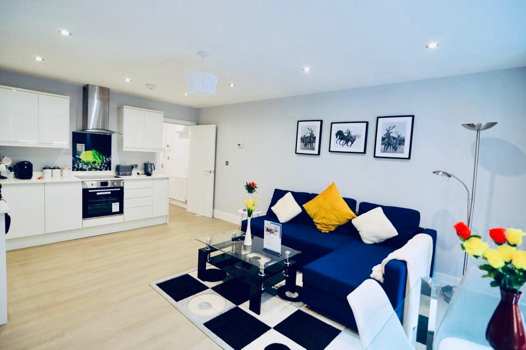 A modern living area features a dark blue sofa with a yellow cushion, paired with a glass coffee table. Black and white artwork adorns the walls. The kitchen is visible, showcasing sleek white cabinets and stainless steel appliances. A dining table with chairs is positioned nearby.