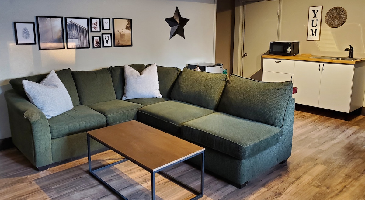 A spacious living area features a large green sectional sofa with decorative pillows. A wooden coffee table is centered in front of the sofa. The wall displays framed pictures, and a kitchenette with a microwave and sink is visible in the background.