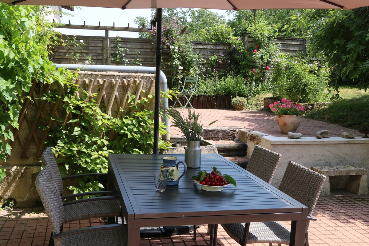 A shaded outdoor dining area features a rectangular table with four chairs, complemented by potted plants and a small blue chair in the background. Lush greenery surrounds the area, along with flower beds and a stone pathway leading to a terraced garden.
