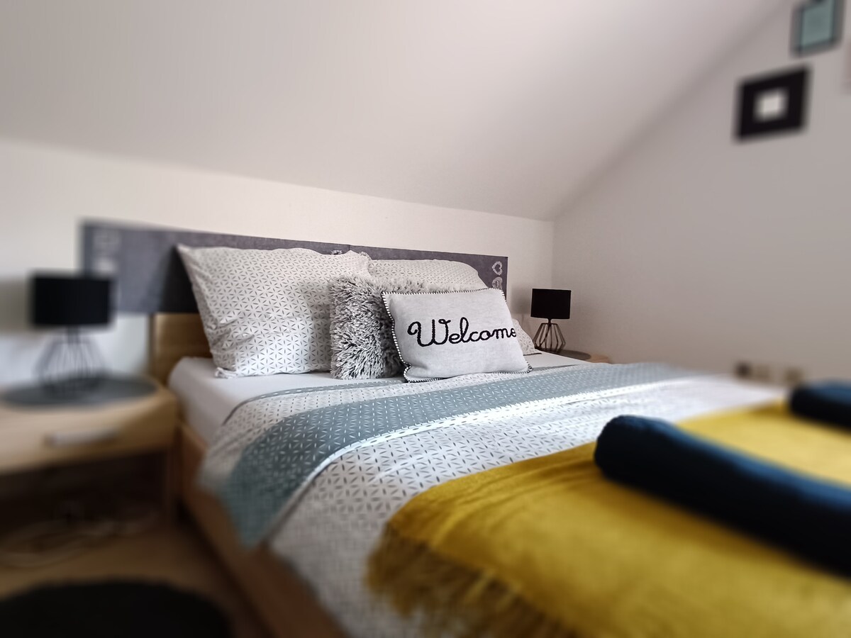 A spacious bedroom features a welcoming atmosphere with a large bed dressed in soft linens. Two bedside lamps provide gentle lighting, and a decorative pillow with the word 'Welcome' adds a friendly touch. A cozy throw in yellow is neatly placed at the foot of the bed.