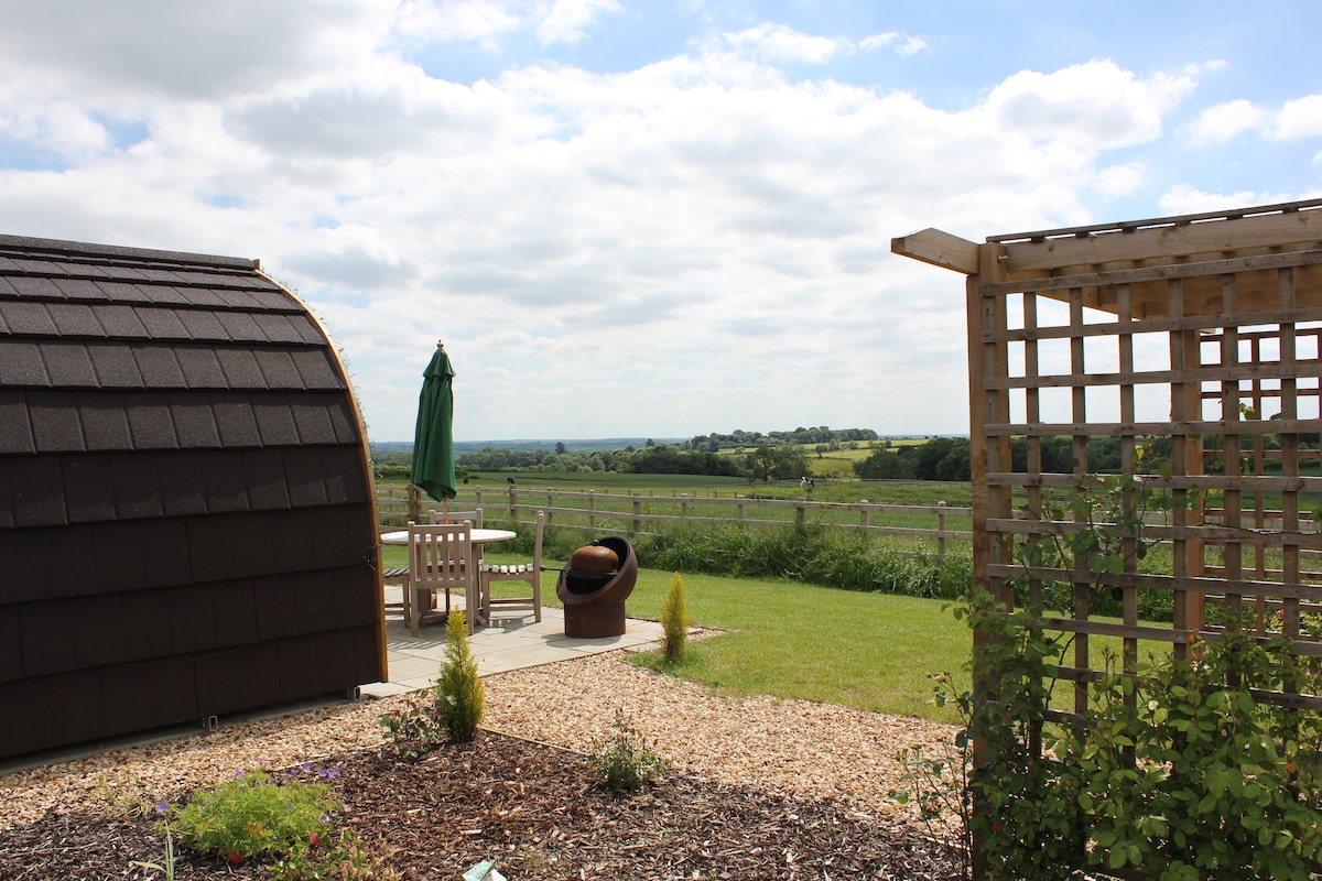 A glamping pod is set against a backdrop of rolling hills, featuring a shaded seating area with a green umbrella and wooden chairs. A fire pit is visible nearby, surrounded by gravel and greenery, creating a relaxing outdoor space.