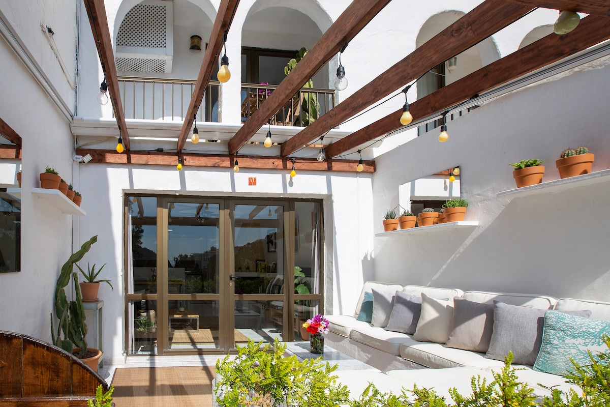 A spacious terrace is adorned with wooden beams and potted plants, creating a serene atmosphere. Comfortable seating is arranged for relaxation, while large glass doors provide views into the home. Soft lighting fixtures add a warm touch to the inviting space.