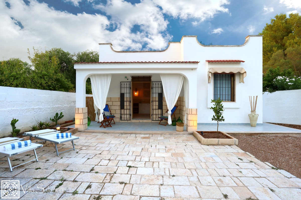 A charming villa exterior is framed by greenery, featuring a stone-paved area with lounge chairs and a rustic wooden table. White curtains gently drape over an entrance, while the sunlight casts shadows on the spacious patio. The inviting atmosphere reflects a serene retreat.