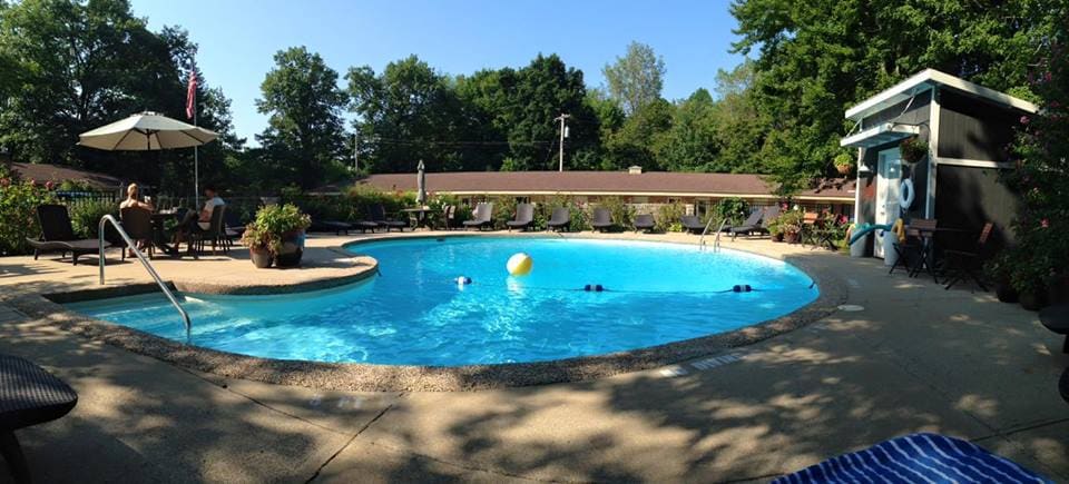 A tranquil pool area is surrounded by lush greenery, featuring lounge chairs and shaded seating under umbrellas. The inviting water reflects sunlight, while a beach ball floats gently. Guests can be seen enjoying the space, creating a relaxed atmosphere in the park-like setting.