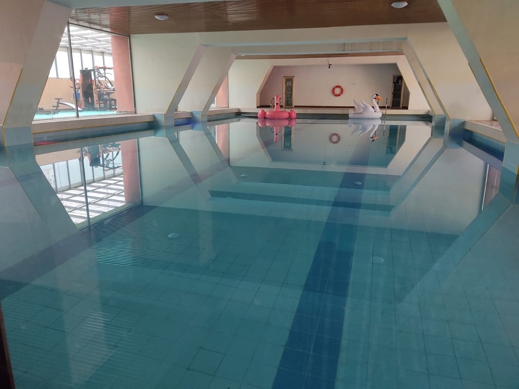 An indoor swimming pool is visible, featuring clear, tranquil water that reflects the surrounding architectural lines. Natural light floods the space through large windows, enhancing the serene atmosphere. Lifebuoys are positioned visibly for safety, while lounging areas can be seen in the background.