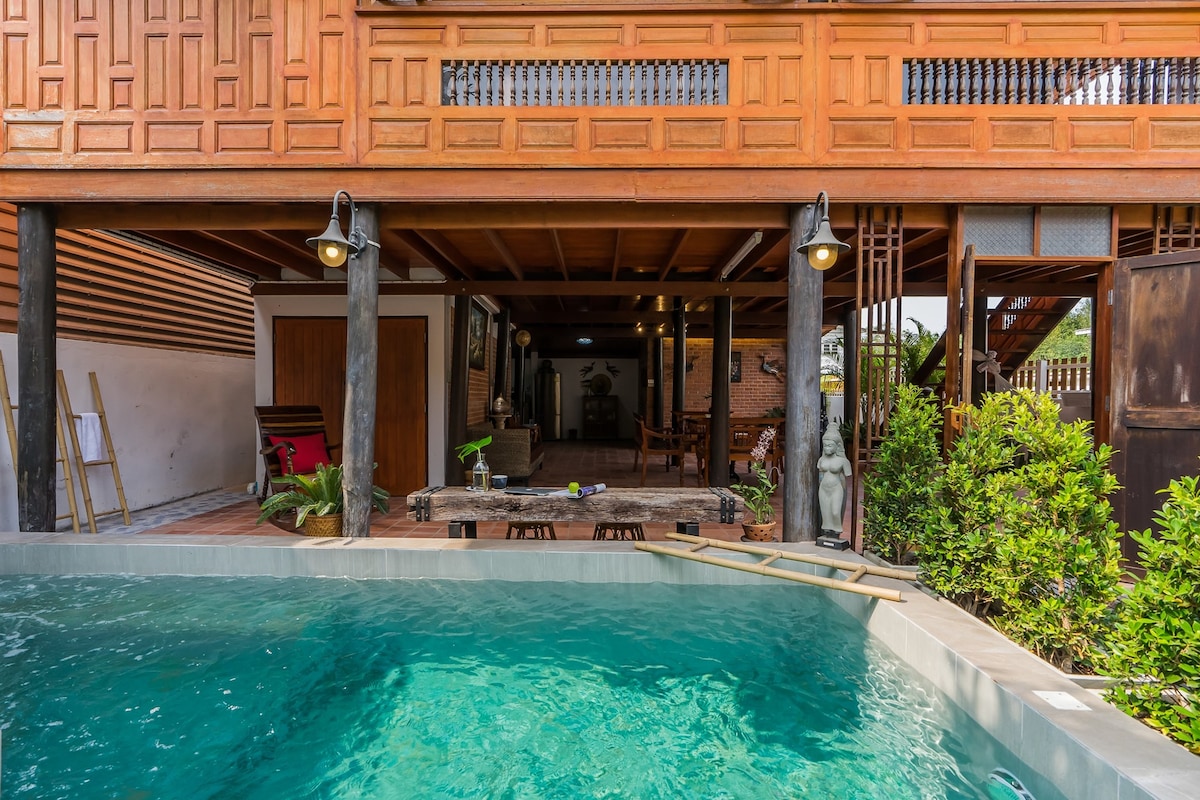 A private outdoor pool is set against a backdrop of a traditional Thai wooden house, featuring an open-air living area beneath a wooden roof. The space is accented by lush greenery and decorative elements, inviting relaxation and leisure.