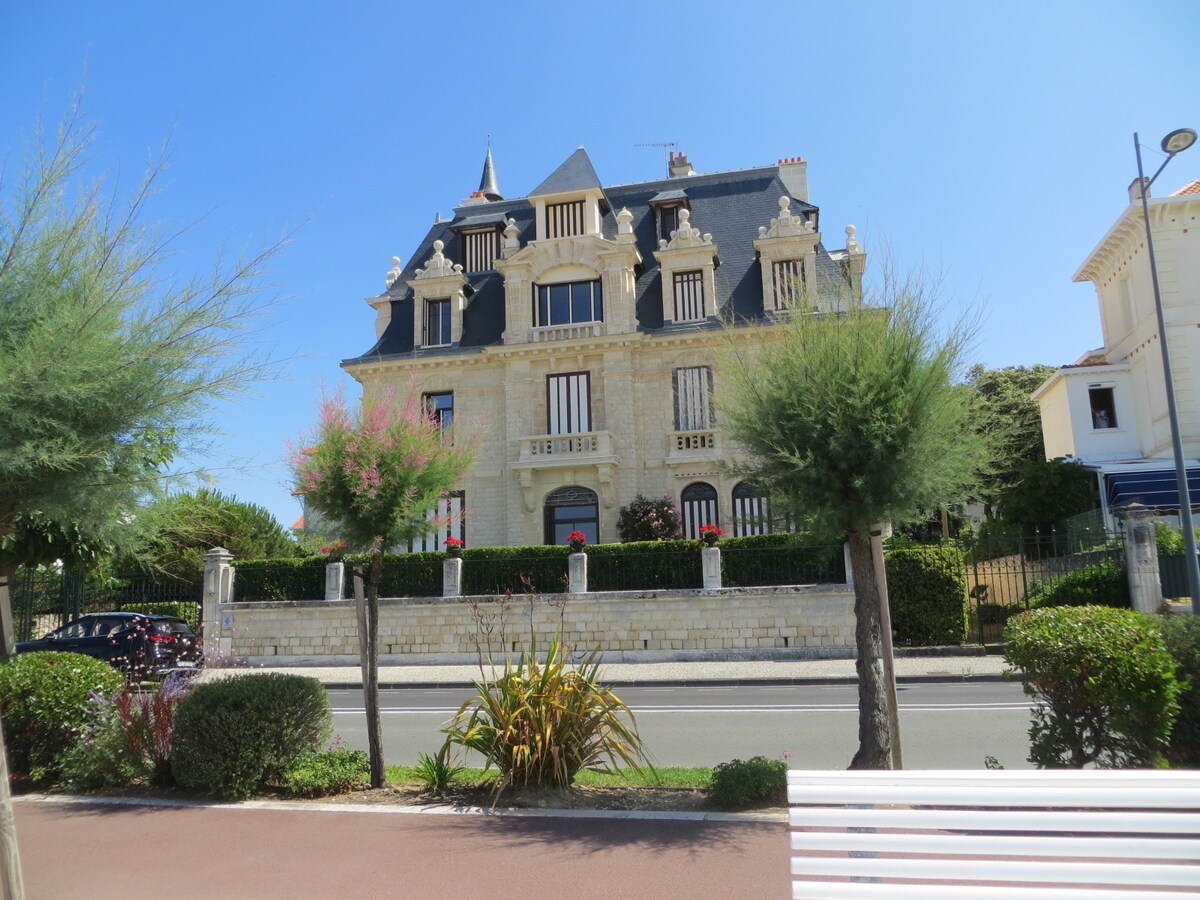 A historical villa, built in 1903, is depicted with a striking facade featuring multiple balconies and prominent architectural details. The building is framed by lush greenery and landscaping, providing a picturesque setting. The clear blue sky complements the villa's elegant structure.