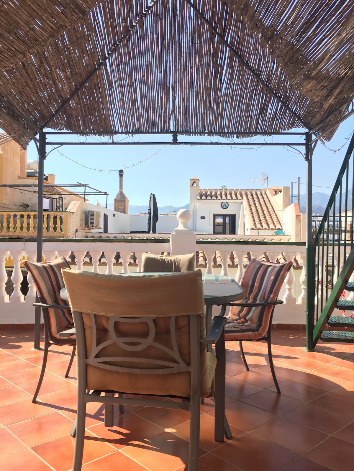 A terrace is seen with a dining table and chairs, shaded by a straw canopy. The surrounding railing offers a view of nearby rooftops and hills. An umbrella stands nearby, adding to the outdoor setting, with bright sunlight illuminating the scene.