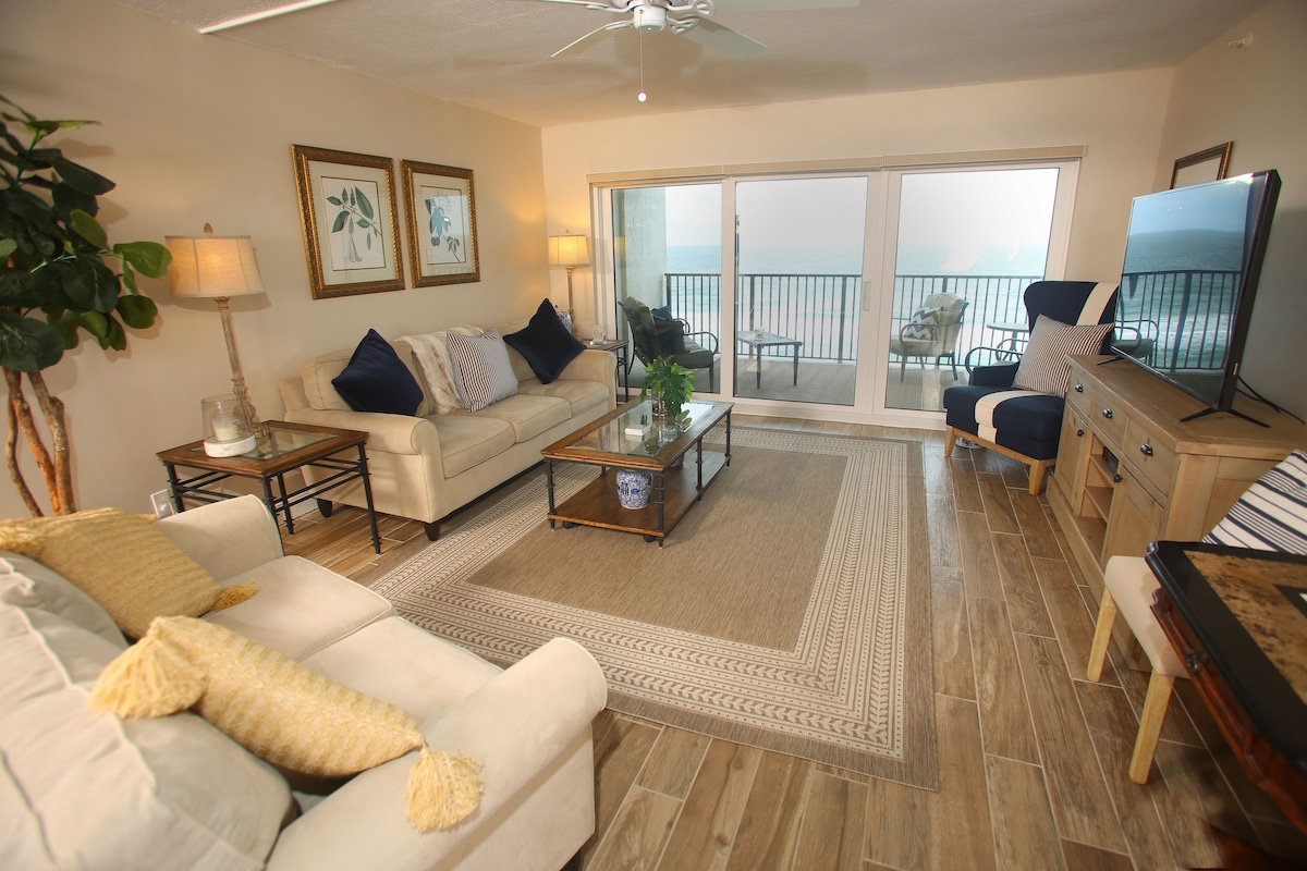 A welcoming living area features soft beige sofas adorned with blue and white pillows, complemented by a wooden coffee table. Large glass doors open to the ocean view balcony, while a coastal-themed rug lies beneath. A flat-screen TV is positioned near the window, enhancing the room's comfort.