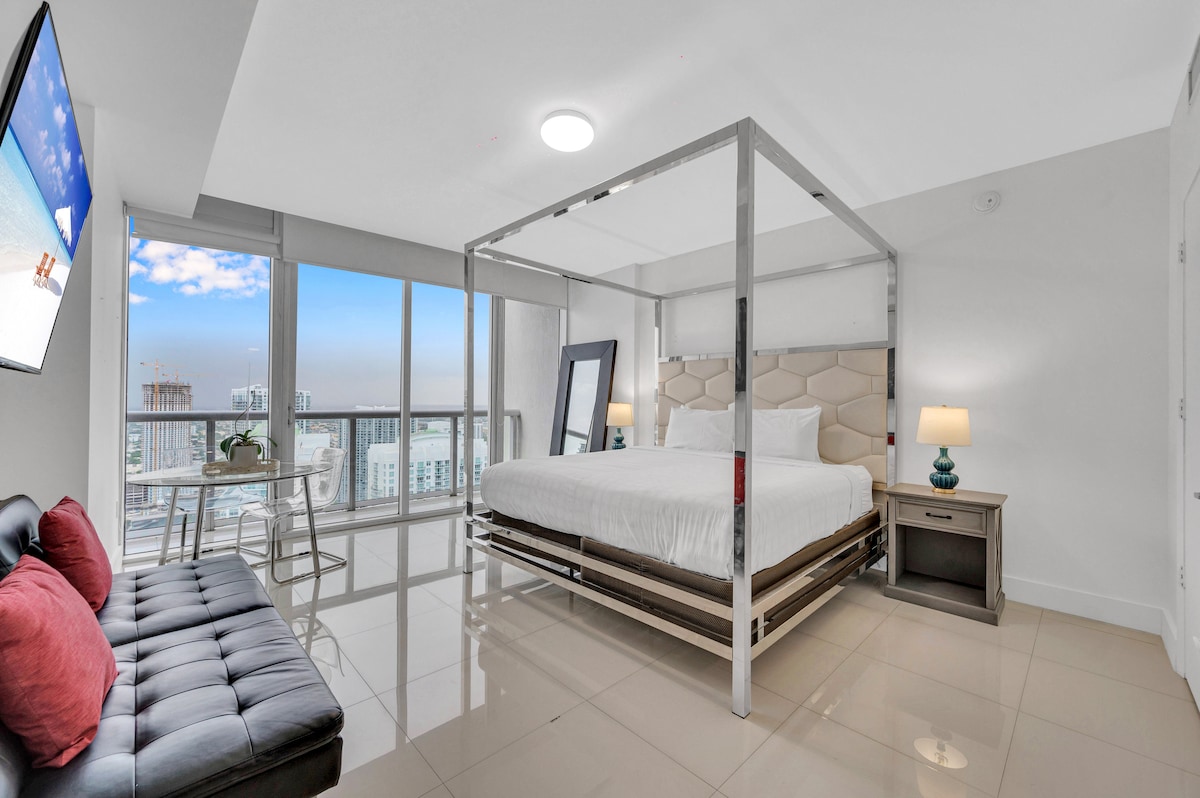 A spacious sleeping area features a stylish four-poster bed with crisp linens and subtle lighting. A comfortable seating area with cushions is visible, alongside a small dining table. Large windows provide a view of the city skyline, allowing natural light to fill the room.
