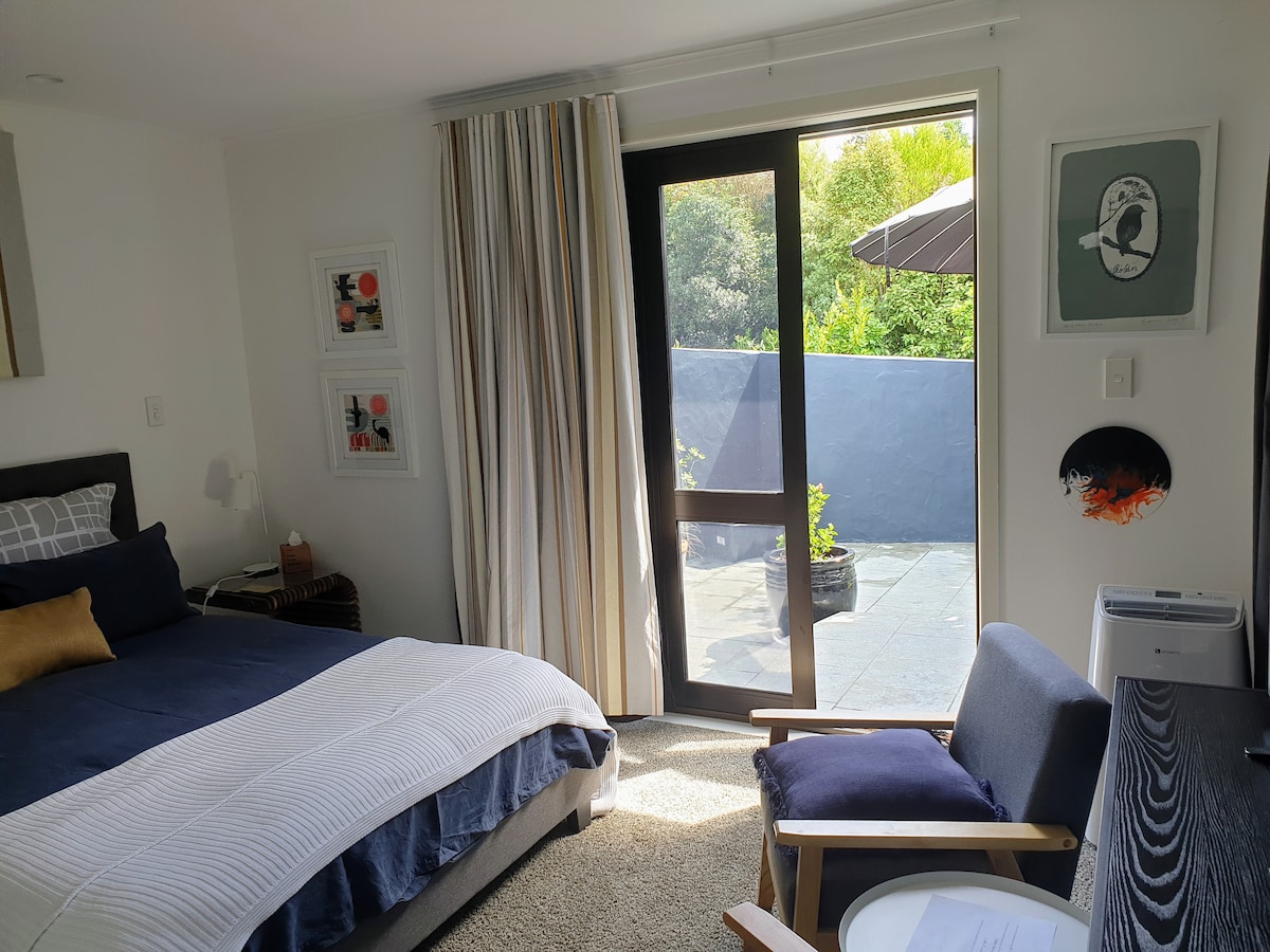 The bright bedroom features a spacious bed with crisp linens and a decorative pillow. A chair is positioned beside a small table. Large glass doors open to a private outdoor area. Soft natural light enhances the room's inviting ambiance.