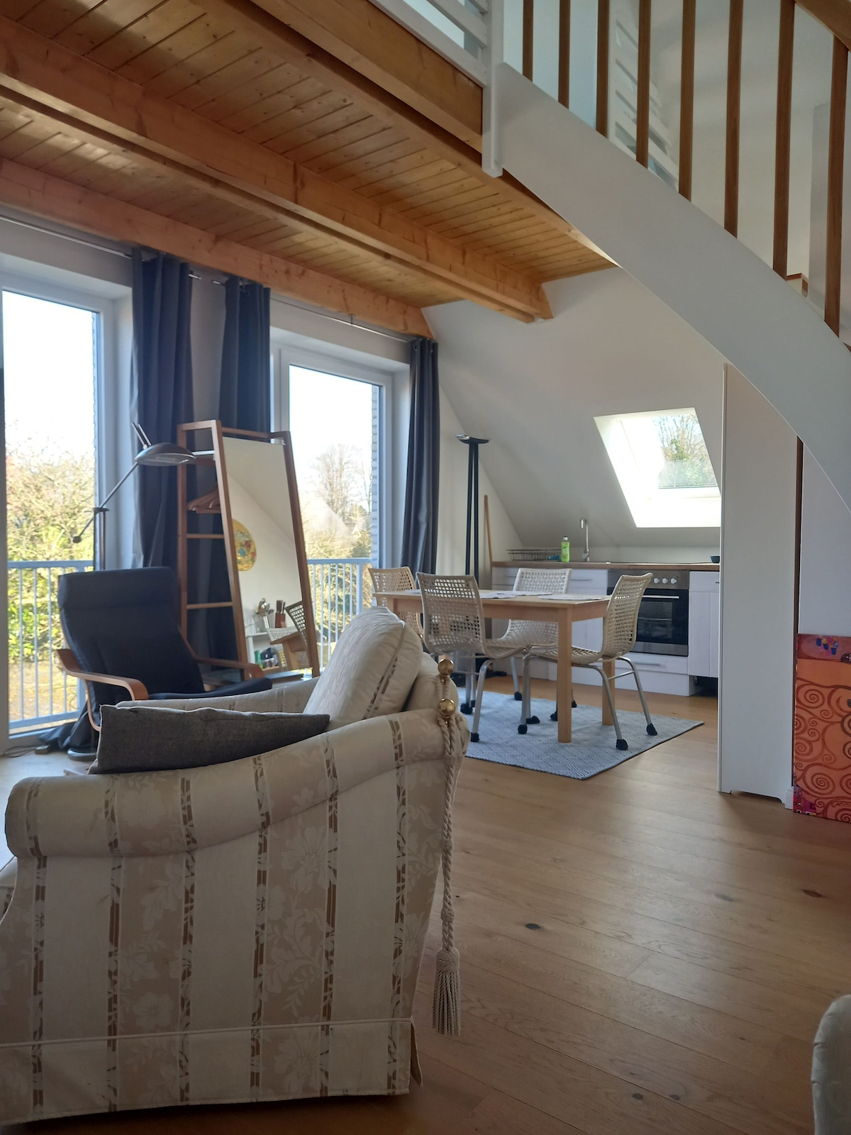 A spacious living area is highlighted by large windows allowing natural light. A comfortable couch and stylish chair are positioned for relaxation. An adjacent dining area features a table surrounded by chairs, with a modern kitchen visible in the background. Wooden beams enhance the ceiling's architecture.
