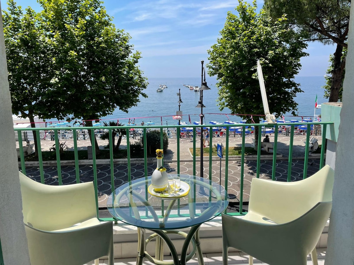A balcony offers a view of the beach and ocean, with sun loungers and umbrellas lining the shore. A glass table is set with a bottle and two glasses, accompanied by two chairs. Lush greenery can be seen framing the scene.
