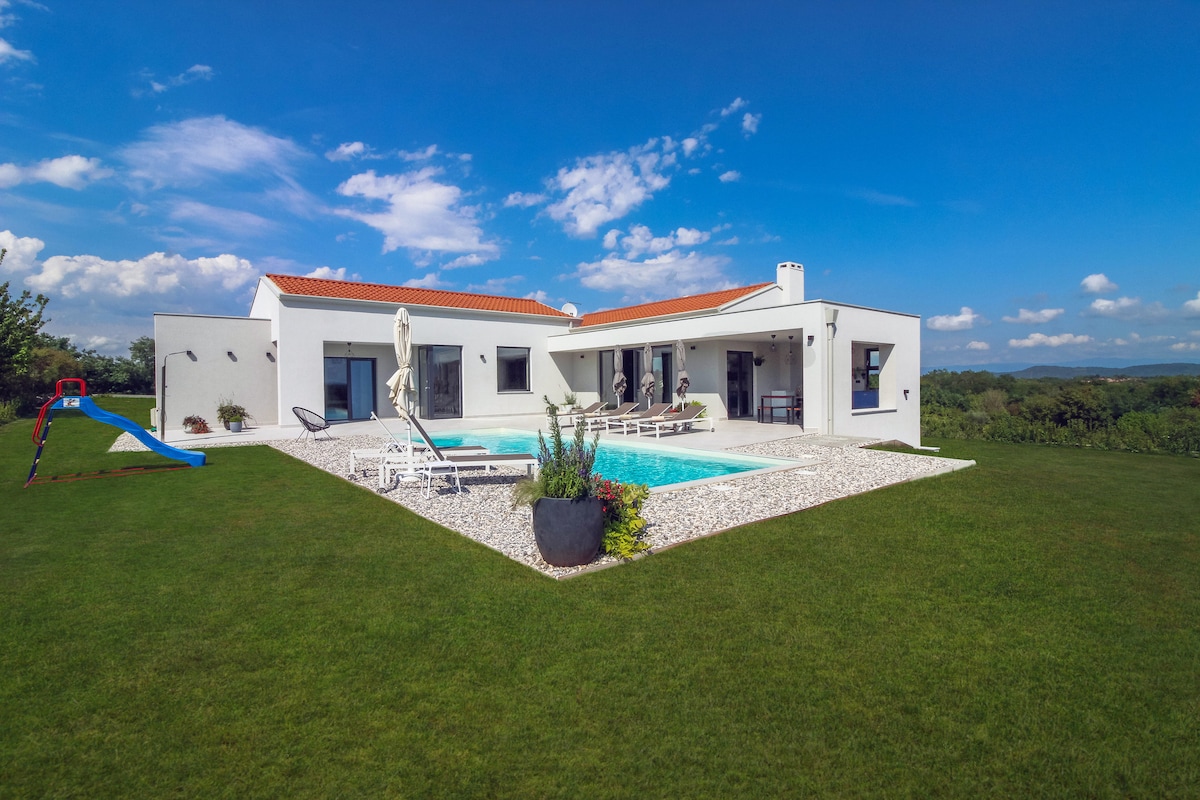 A modern villa is showcased against a clear blue sky, featuring a spacious outdoor area with a glimmering pool. The property is surrounded by lush green grass, complemented by lounge chairs and playful elements like a small slide. Scenic hills are visible in the background.