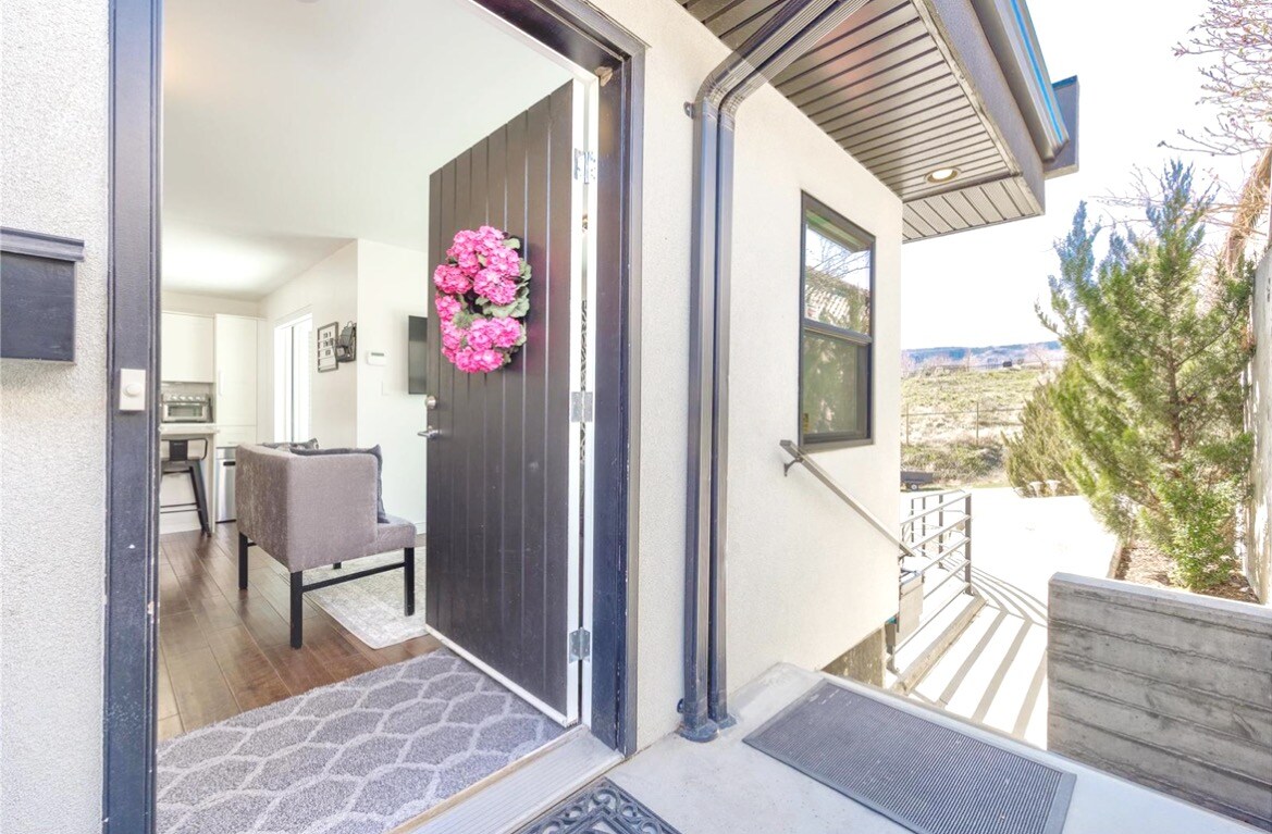 A welcoming entrance features a dark door adorned with a floral wreath. Light is captured from the adjacent living area visible through the open door. A gray rug lies on the floor, leading to an outdoor space with landscaped greenery.