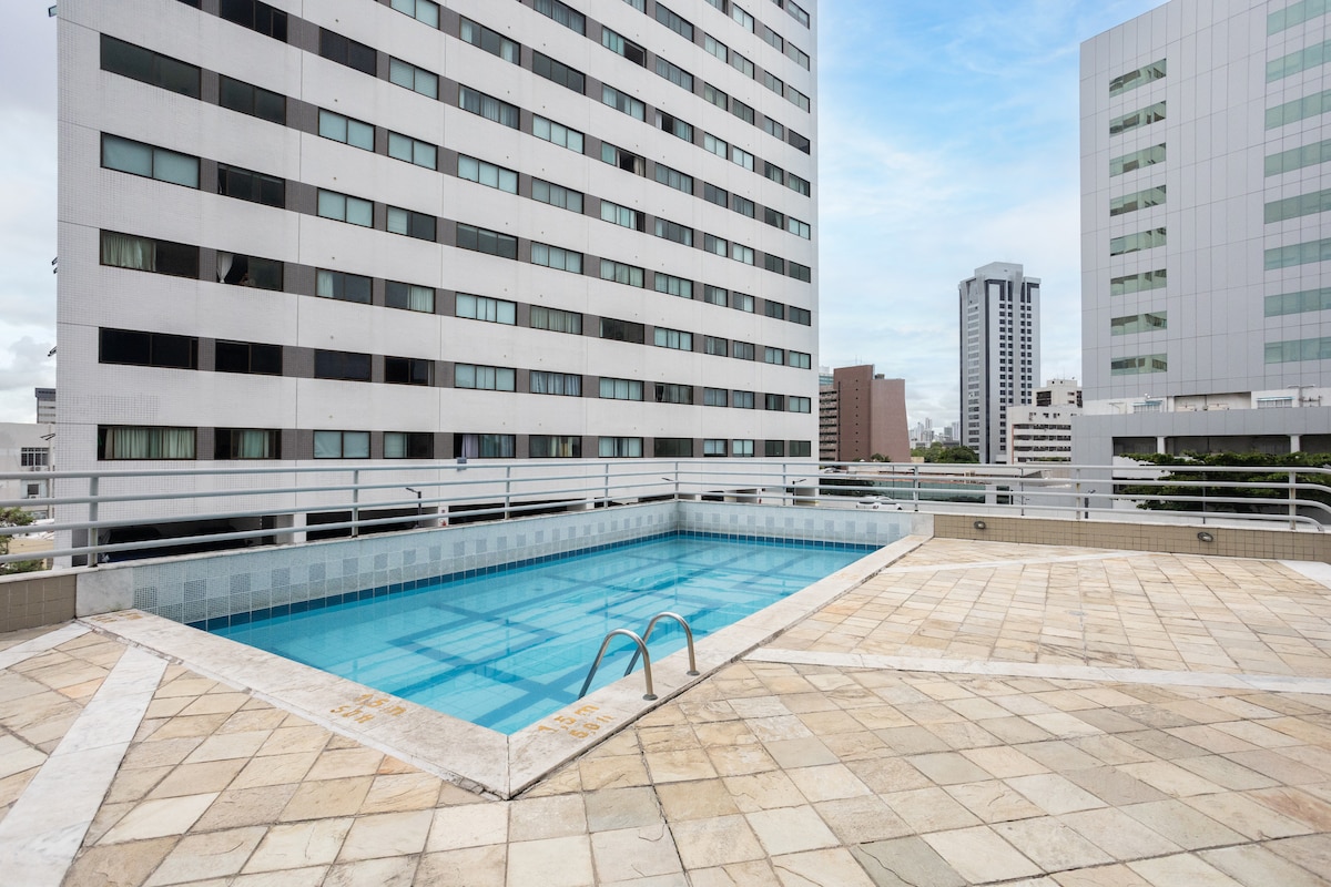 An outdoor swimming pool is available, surrounded by tiled flooring and featuring a gentle gradient from shallow to deep. The poolside area is bordered by tall buildings, offering a glimpse of the city skyline in the background, with clear skies above.