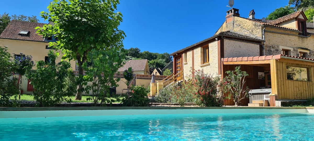 A clear blue pool reflects the bright sky, surrounded by landscaped gardens. The charming house features warm stone walls and wooden accents, alongside a pool house. Lush greenery and trees enhance the outdoor space, providing a peaceful atmosphere for relaxation.