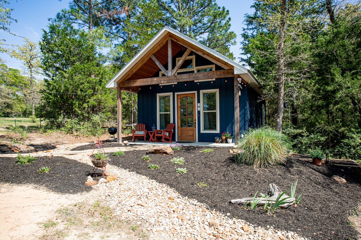 A charming cottage exterior is framed by native trees and landscaped with decorative stone pathways. The deep blue siding contrasts with natural wood accents and features large front doors flanked by windows. Two red Adirondack chairs are positioned on the porch, inviting relaxation.