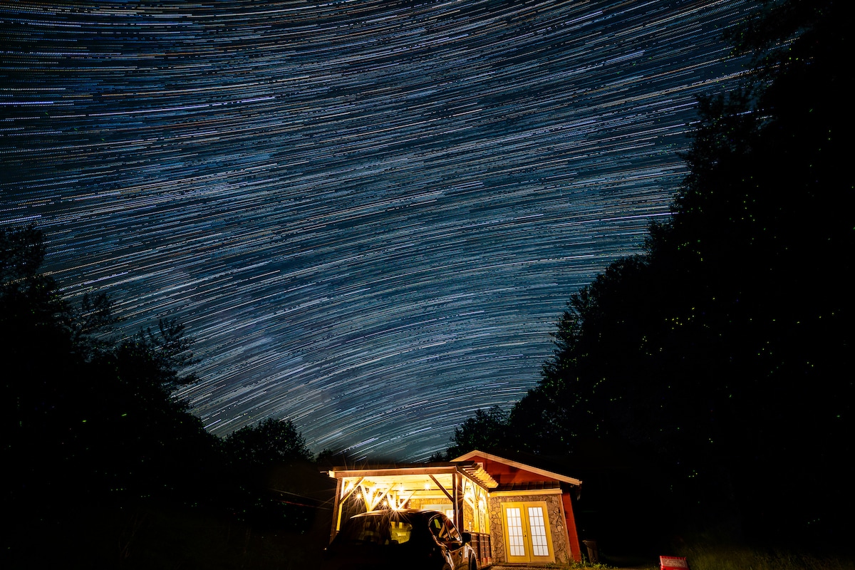 A cozy guesthouse is illuminated under a starry night sky, with trailing stars creating a dynamic backdrop. The warm glow from the windows adds a welcoming touch, while the surrounding trees provide a natural frame to the scene.