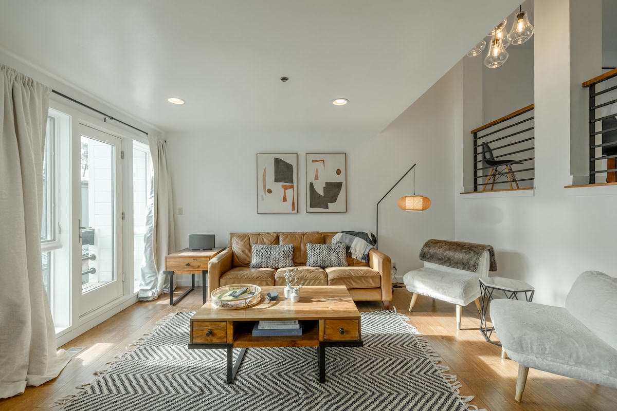 A modern living area features a spacious leather sofa adorned with throw pillows, complemented by a wooden coffee table. Two abstract art pieces hang on the wall. Natural light enters through double doors, illuminating the room's wooden floor and neutral decor.