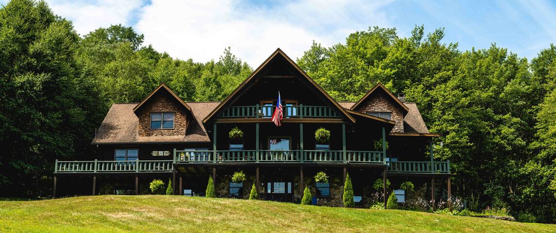 The exterior of a large lodge is viewed from a grassy area, showcasing a wide porch with planters and an American flag. Surrounded by trees, the structure features a combination of stone and wood elements, providing a rustic aesthetic under a clear blue sky.